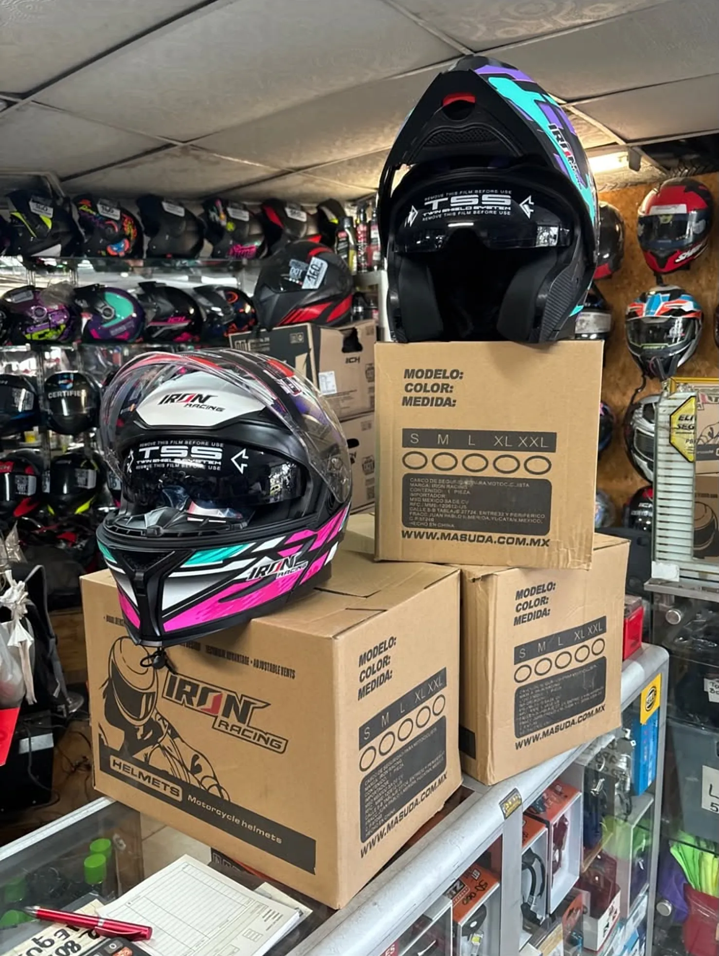 Cascos Iron Racing nuevos sobre sus cajas en el mostrador, con la pared de cascos detrás